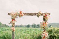 a colorful rustic wedding arch of rough wooden slabs and white and coral blooms plus a backdrop of a green field