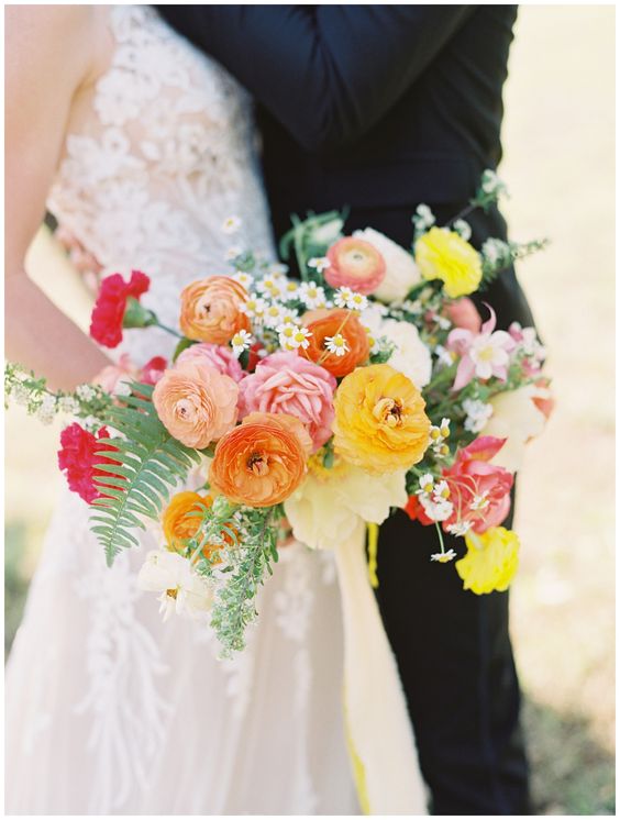 a colorful wedding bouquet including orange, blush and yellow raunuculus, greenery and some chamomiles is creative