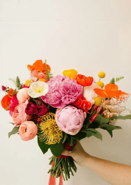 a colorful wedding bouquet with red poppies, peachy and yellow ranunculus, pink peonies, pincushion proteas and foliage