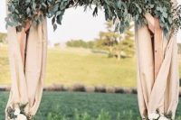a cool rustic wedding arch covered with neutral burlapm eucalyptus, white floral arrangements attached is a chic decoration