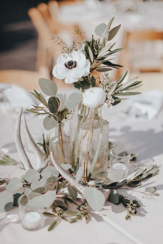 a creative wedding centerpiece of mismatching bottles, greenery, a white ranunculus and a white aemone, antlers and candles around