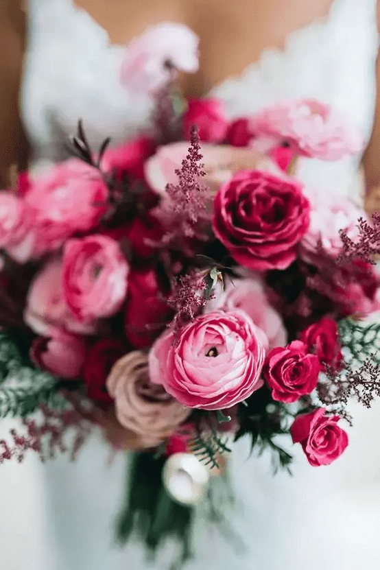 A jewel tone wedding bouquet of pink ranunculus and fuchsia roses, some dark and usual foliage is amazing for fall