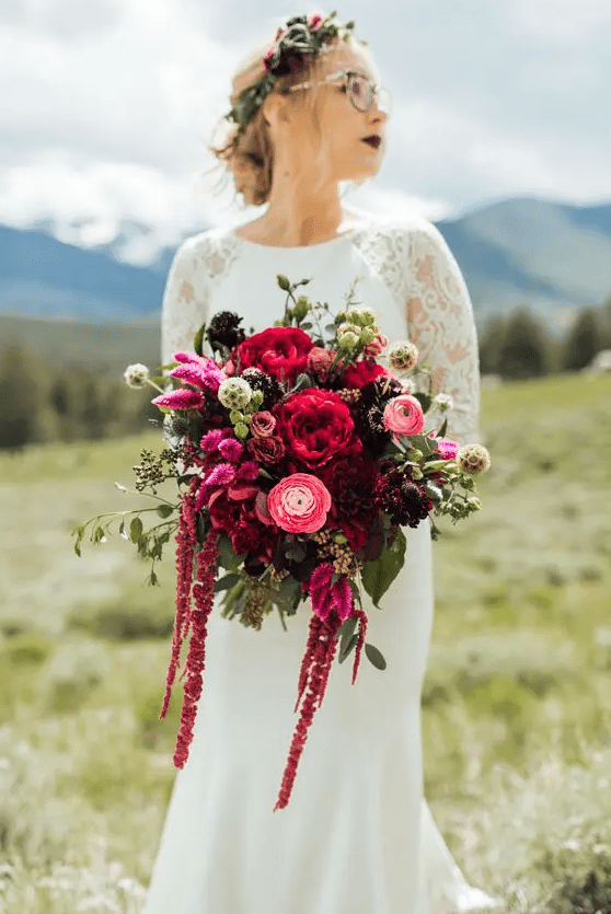 A jewel tone wedding bouquet of pink, red and burgundy blooms including ranunculus, peonies, amaranthus and some greenery and seed pods