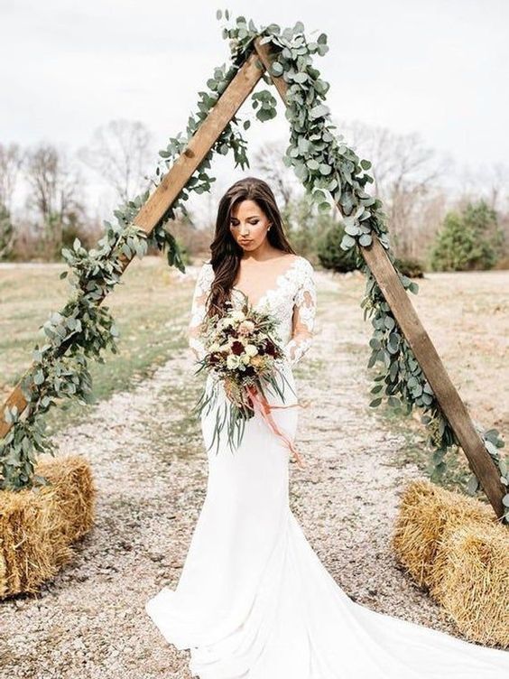 a pretty rustic triangle wedding arch covered with greenery and with hay at the base is a cool solution for a rustic boho wedding
