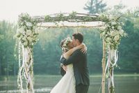 a pretty rustic wedding arch of brich branches, with white ribbons, white blooms and greenery and a cool backdrop of a pond
