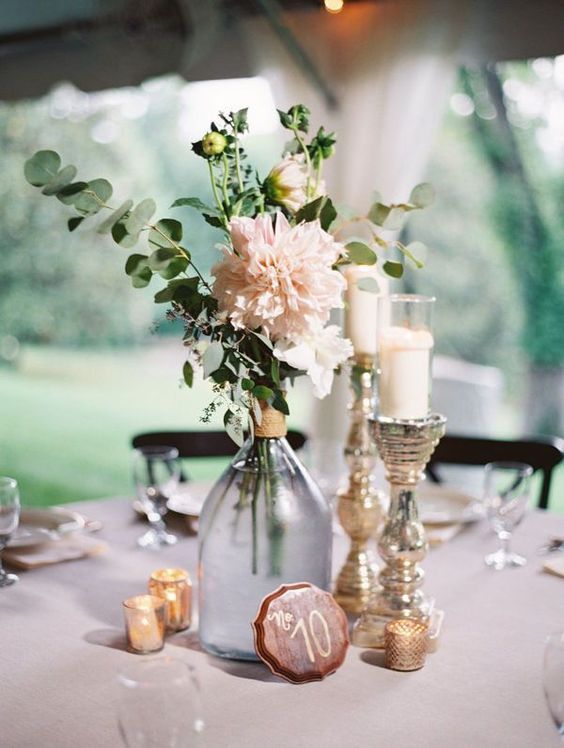 a pretty wedding centerpiece of pink dahlias, greenery, candles, a table number and mercury glass candleholders