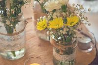 a relaxed rustic wedding centerpiece of a wood slice, candles, jars wrapped with lace, baby’s breath, yellow daisies and white carnations