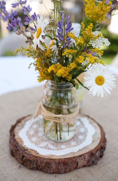 a relaxed rustic wedding centerpiece that includes chamomiles and wildflowers, a jar placed on a wood slice and a doily