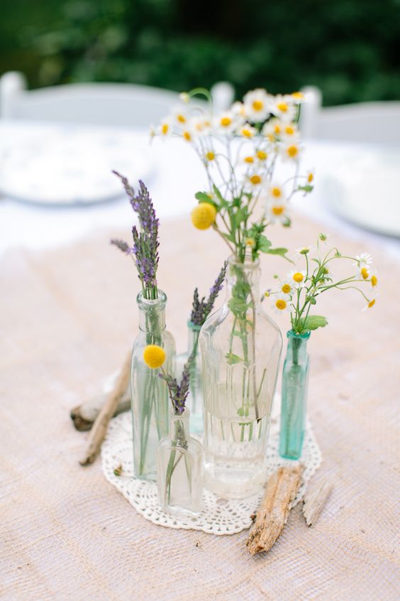 a relaxed wedding centerpiece of bottles and vases with lavender and daisies, billy balls and driftwood for a boho wedding