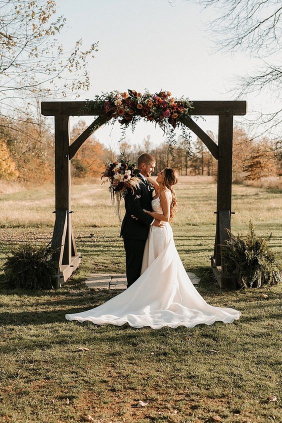 a rustic wedding arch decorated with greenery, blush and dark blooms and dark greenery arrangements around