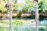 a rustic wedding arch of birch branches, pastel blooms and greenery with a backdrop of a pond is a lvoely idea for summer