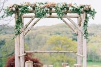 a rustic wedding arch with tree stumps, greenery and peachy blooms plus a view of a fall forest for an autumn rustic wedding