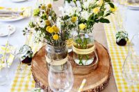 a rustic wedding centerpiece of a wood slice with jars, billy balls, daisies and yellow garden roses is extremely cute and bright