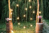 a simple rustic wedding arch of wooden branches, some baby’s breath, lights, pink floral arrangements on barrels next to the arch