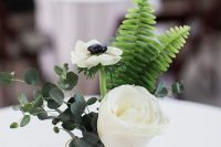 a stylish and simple wedding centerpiece of a vase with a white rose and ranunculus, some eucalyptus and fern and candles around