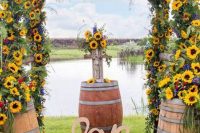 a super colorful summer rustic wedding arch covered with greenery, sunflowers, bold red and blue flowers and barrels at the base