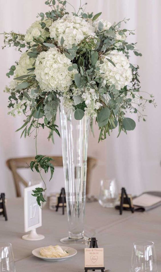 a tall wedding centerpiece of a sheer vase, white hydrangeas and various greenery and eucalyptus is chic and timeless