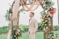 an incredibly romantic wedding arch or rattan, with greenery and fern, blush, orange and hot red blooms and with some blush fabric is chic