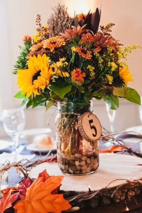 a bright and cute rustic fall wedding centerpiece of a jar with pebbles, orange, rust, yellow and deep purple blooms and leaves