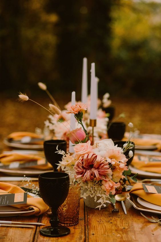 a beautiful fall wedding tablescape with pink blooms and greenery, black and amber glasses, tall candles and mustard napkins