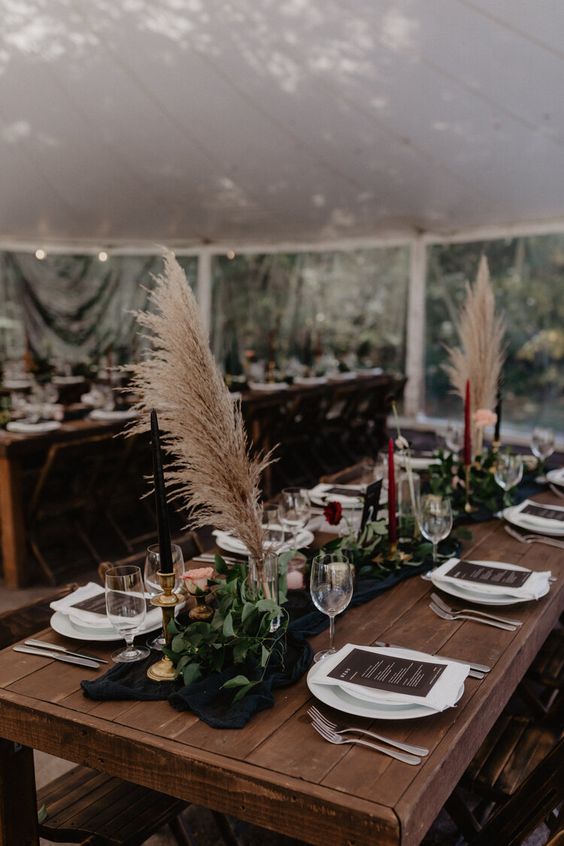 a beautiful moody fall wedding tablescape with a black runner and greenery, pampas grass and black and burgundy candles, gold touches and dark menus