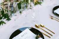 14 a modern elegant wedding tablescape with white linens, black plates, gold cutlery, a greenery runner with candles and neutral blooms
