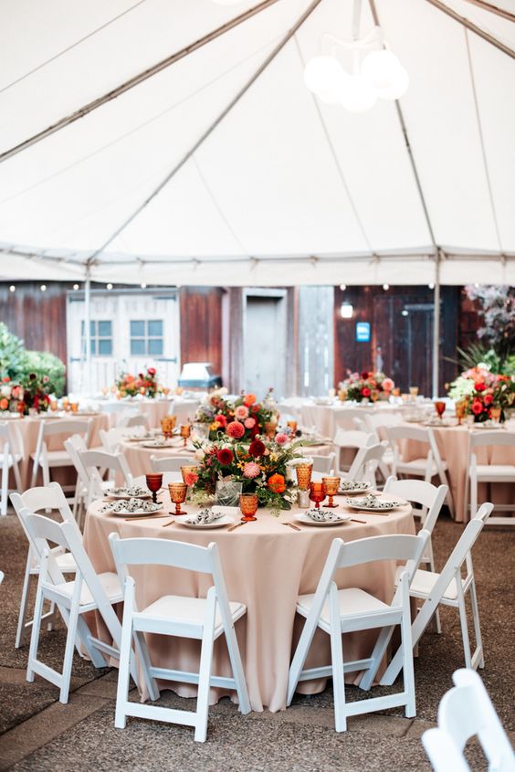 a bold fall wedding reception space with neutral linens, bold orange, burgundy and pink blooms and greenery and amber glasses