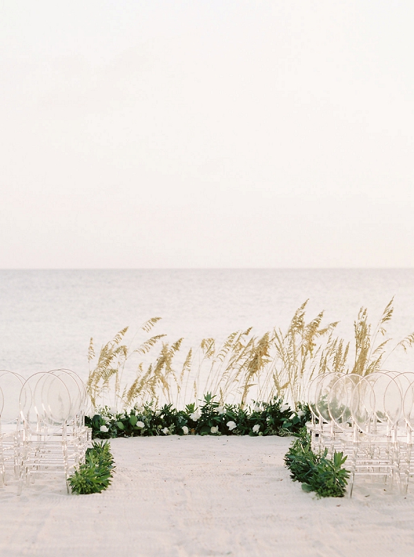 a modern beach wedding ceremony space with overgrown grasses, greenery, white blooms, ghost chairs and greenery lining up the aisle