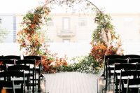 16 a modern fall wedding ceremony space with a round wedding arch decorated with greenery and bright fall leaves, with candle lanterns lining up the aisle