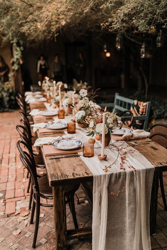 a relaxed rustic fall wedding tablescape with a neutral runner, amber glasses, terracotta vases with neutral and pastel blooms, candles and menus