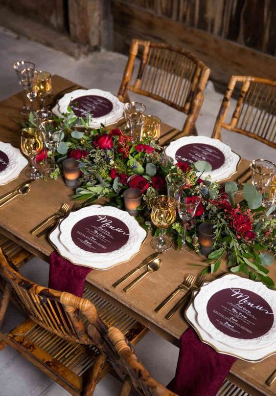 a very elegant and refined rustic fall wedding tablescape with a greenery and red rose runner, candles, burgundy menus and napkins plus gold cutlery