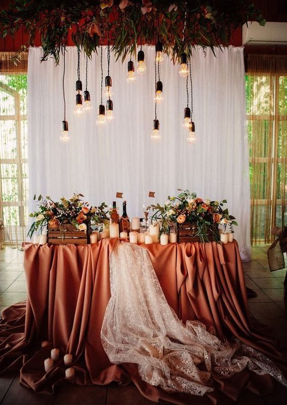a fabulous rustic wedding reception space with a rust colored tablecloth and a lace runner, bold blooms and greenery in crates and candles plus bulbs over the space