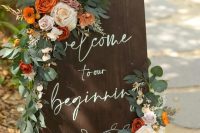 a rustic fall wedding sign with greenery, white, rust, orange and blush blooms and calligraphy is a stylish and cool idea