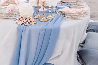 a beach wedding tablescape with a blue runner, pink napkins, a cake topped with pink blooms, driftwood and some elegant candles