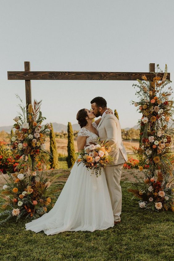 a beautiful and colorful rustic fall wedding arch lushly decorated with greenery, blush, rust, pink, orange and burgundy blooms, greenery and pampas grass