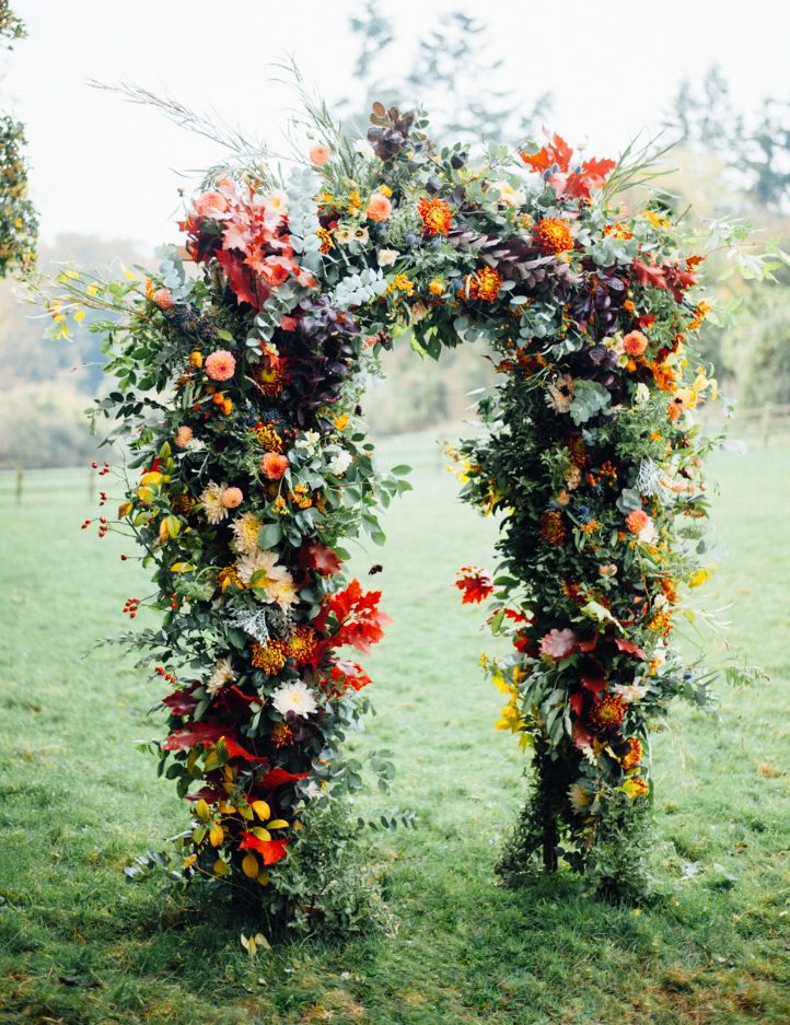 a beautiful and colorful rustic fall wedding arch with greenery, red, yellow, pink blooms, colorful fall leaves is a lovely idea for the season