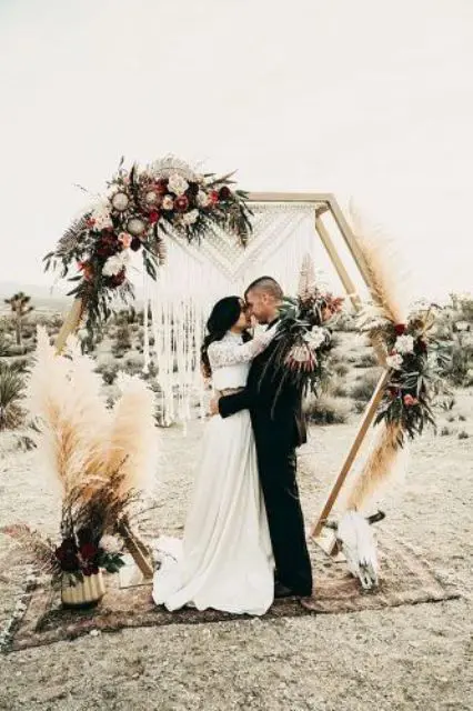 a boho fall hexagon wedding arch decorated with greenery, pampas grass, white, pink and deep purple blooms, macrame and an animal skull