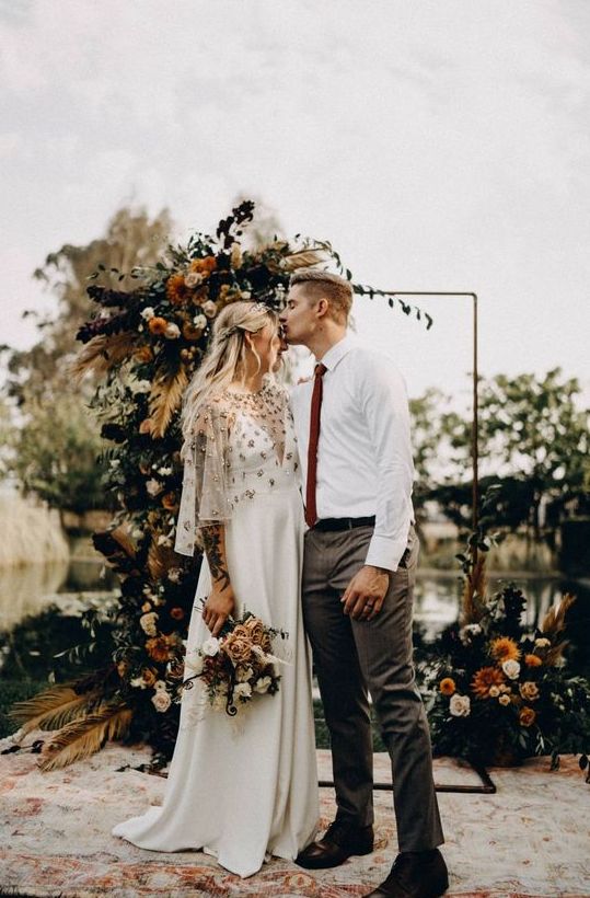 a boho fall wedding arch decorated on one side and at the base with greenery, pampas grass, feathers, bold and dark blooms