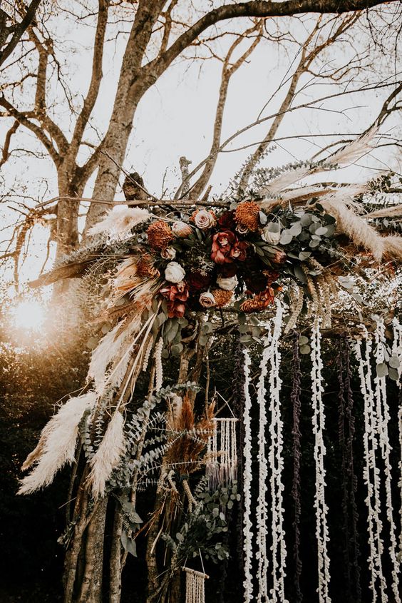 a boho fall wedding arch with greenery, blush, rust and burgundy blooms, pampas grass, tassels and garlands of petals is wow