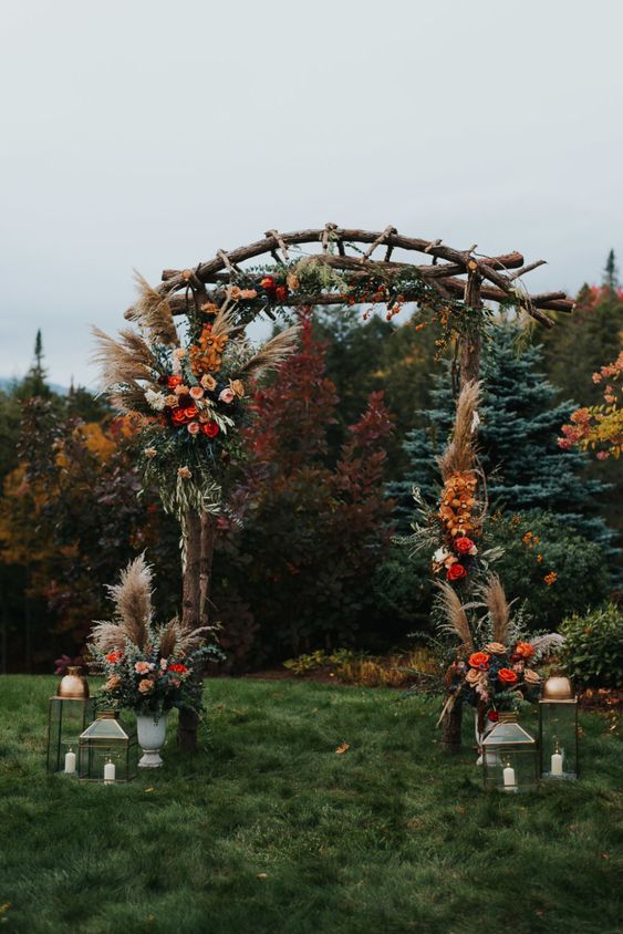 a boho rustic fall wedding arch with pampas grass, pink, blush, red and burgundy blooms, greenery and fall leaves plus candle lanterns