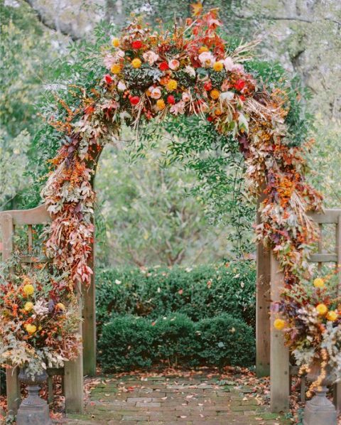 a bold and lush rustic fall wedding arch decorated with greenery, bold foliage, pink, red, mustard and yellow blooms and fruits