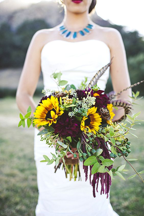 a bold and sumptuous wedding bouquet with lots of textural greenery, dark blooms, feathers and sunflowers is a cool idea for a fall boho bride