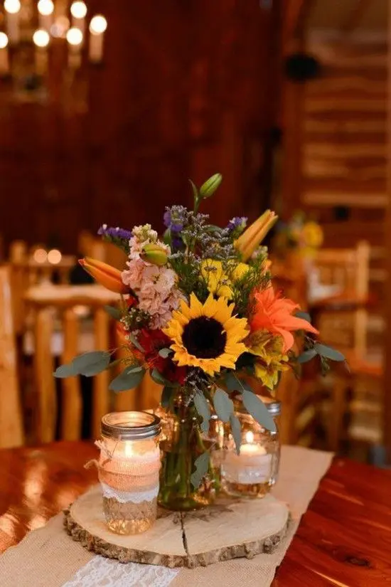 a bold wedding centerpiece of colorful lilies, sunflowers, roses and hydrangeas and candles around is a cool idea for a rustic wedding