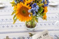 a bright and pretty summer wedding centerpiece of sunflowers and bold blue flowers, mimosas and some greenery is amazing for a colorful wedding