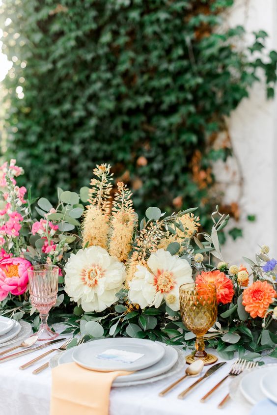 a colorful secret garden wedding tablescape with a greenery and colorful bloom runner, colorful glasses, layered plates and gold cutlery