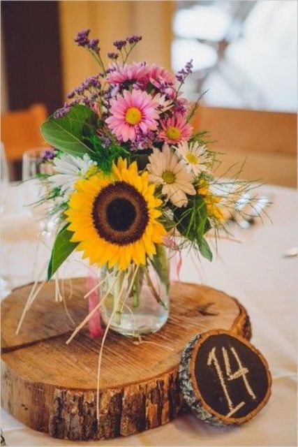 a colorful summer wedding centerpiece of sunflowers, pink and white blooms, greenery and lavender placed on a wood slice and with a wood slice table number