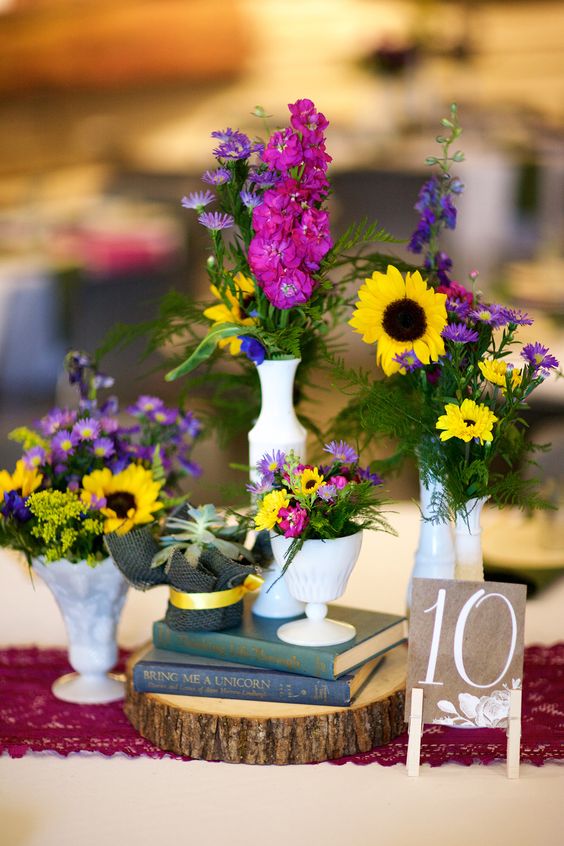 a colorful vintage inspired wedding centerpiece of a wood slice, books, vases with colorful blooms and greenery and a table number