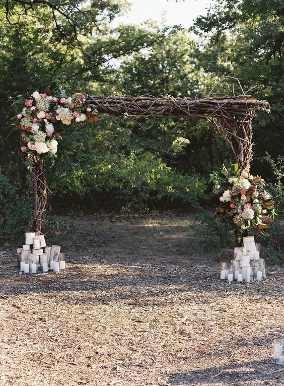 a cool rustic wedding arch of branches, twigs, white, green, pink, deep purple blooms, greenery and lots of candles at the base