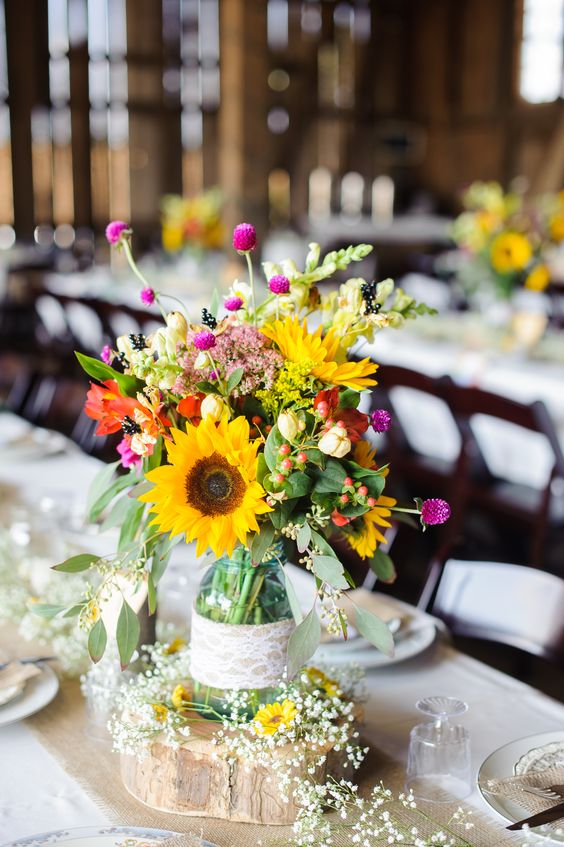 a cute and colorful rustic wedding centerpiece of a jar with sunflowers, red, pink blooms, greenery and berries plus baby's breath around is cool