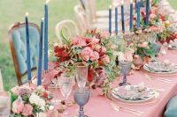 a fairy-tale wedding table setting with blue candles, glasses and candleholders, pink and red floral centerpieces and gold cutlery